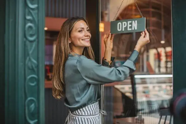 Smiling female small business owner hanging an 'Open' sign at the entrance of her cafe